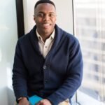 A professional black man smiling while seated by a window, embodying modern business vibes.