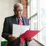 Elderly man in business attire examining papers by a window in an office setting.